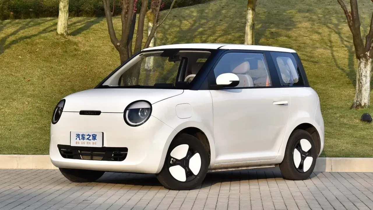Auto elettrica piccola 4 posti, bianca, con tetto nero, parcheggiata su un vialetto pavimentato con alberi sullo sfondo.
