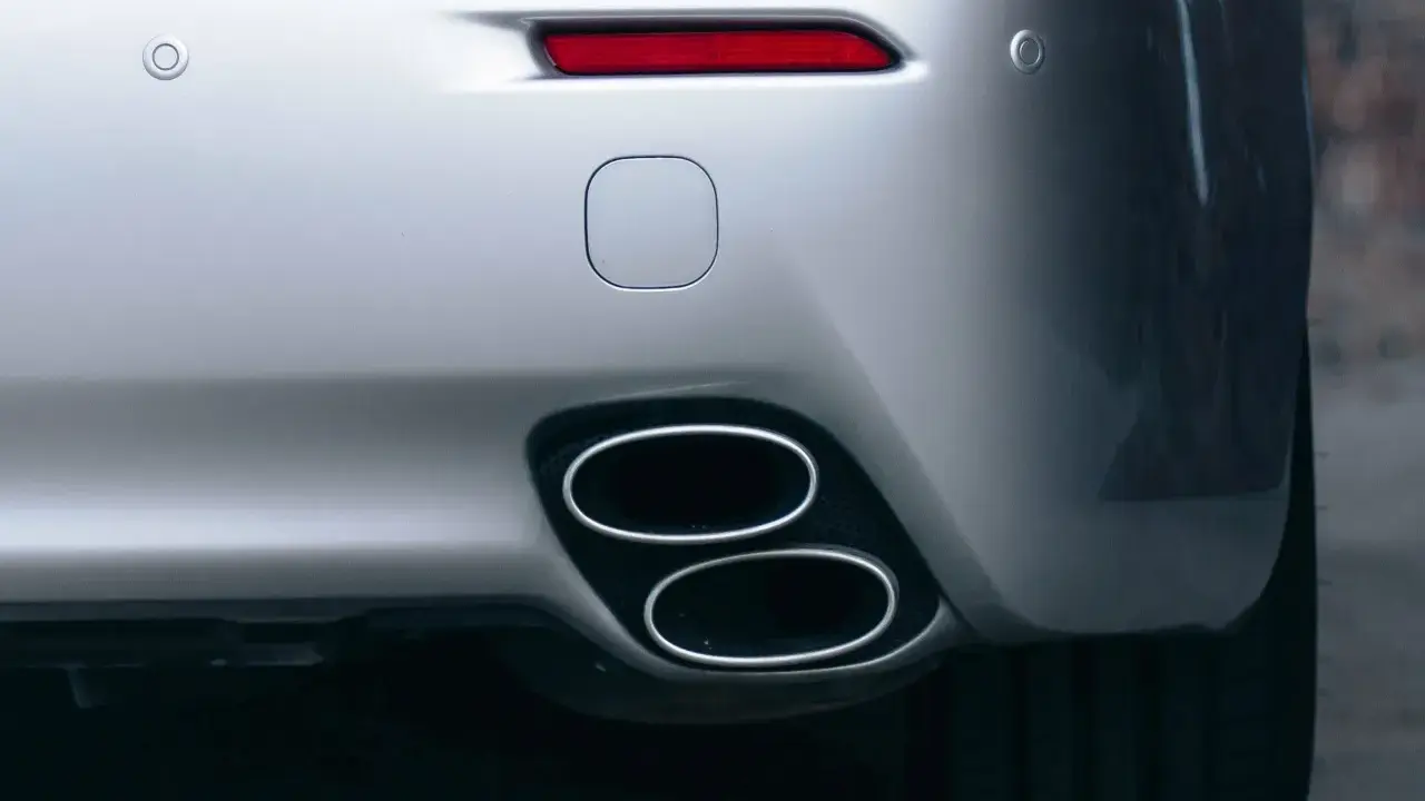 Doppio scarico cromato su un'auto sportiva argentata, parte del collettore di scarico. Sensori di parcheggio e fanale posteriore rosso visibili.