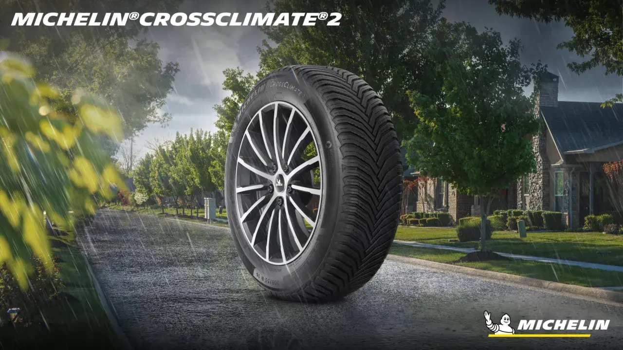 Pneumatico Michelin CrossClimate 2 in primo piano su strada bagnata, con case e alberi sullo sfondo. Nessun difetto visibile.