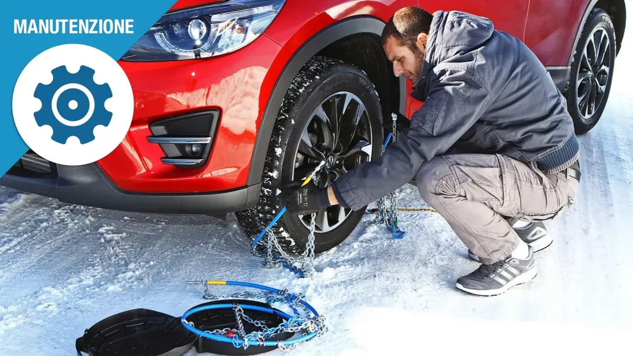 Uomo monta catene da neve su una ruota di un'auto rossa in un paesaggio innevato. Le gomme invernali sono essenziali per la sicurezza.
