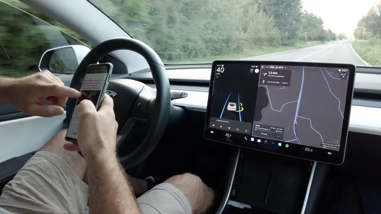 Mani sul volante, ma lo sguardo è sullo smartphone mentre il **Tesla Autopilot** guida. Navigazione e musica sul grande schermo.