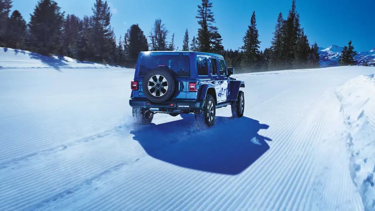 Un blu Jeep Wrangler affronta la neve con pneumatici Toyo Open Country AT3, lasciando tracce su un sentiero battuto tra alberi innevati e montagne.