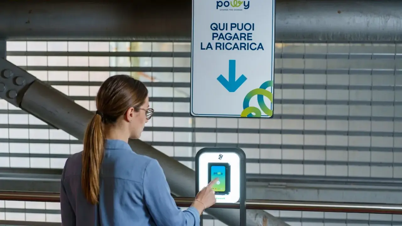 Donna interagisce con un terminale per pagare la ricarica dell'auto elettrica. Il cartello indica "Qui puoi pagare la ricarica", suggerendo il costo ricarica auto elettrica.