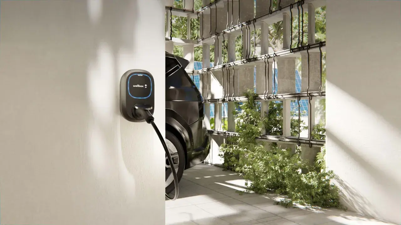 Wallbox per auto elettriche, con cavo collegato a un'auto scura. L'ambiente è moderno, con elementi verdi e pannelli solari visibili.