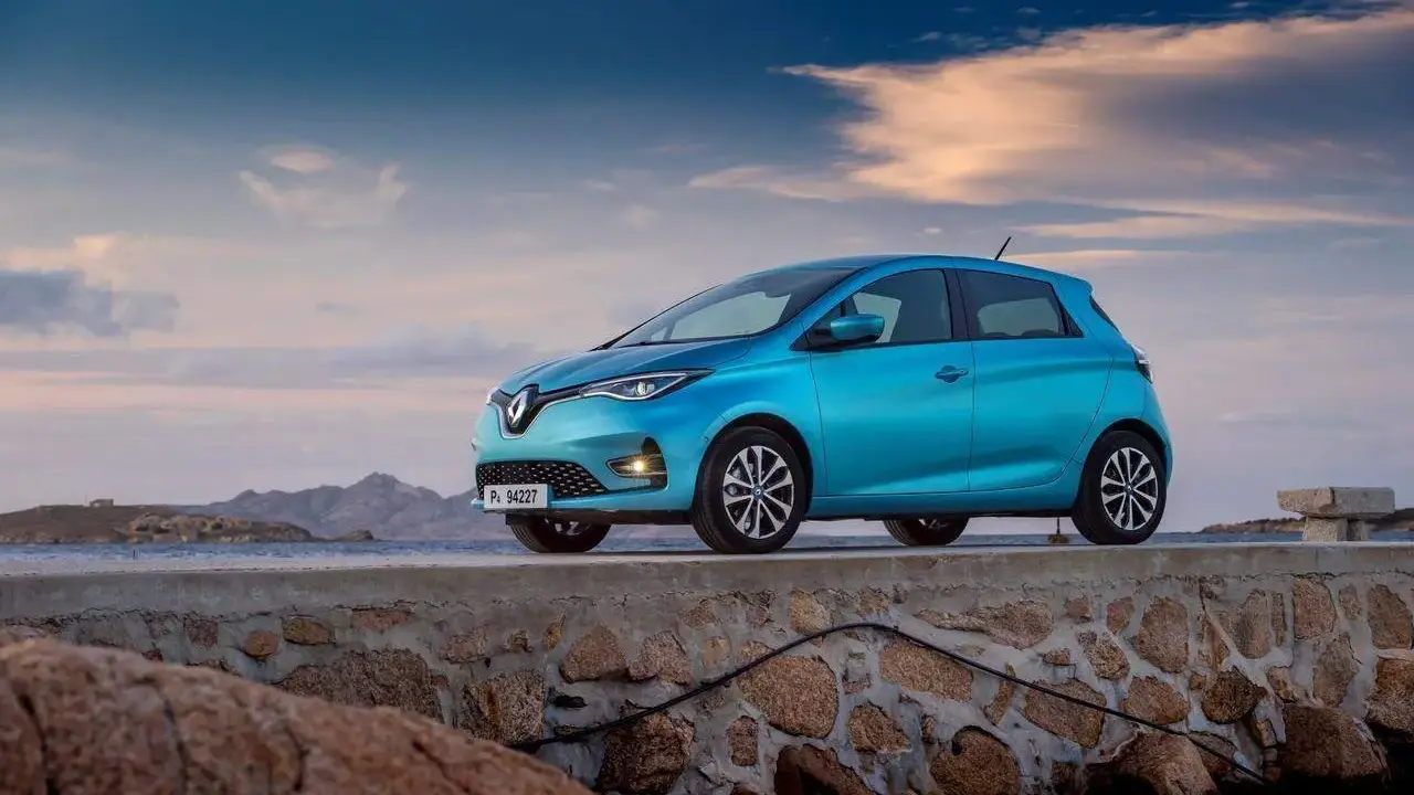 Un'elegante Renault Zoe blu, uno dei tanti modelli Renault, è parcheggiata su un muretto di pietra con il mare e le montagne sullo sfondo.