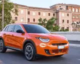 Auto elettrica arancione in movimento, con 400 km di autonomia, su strada urbana con edifici storici sullo sfondo.