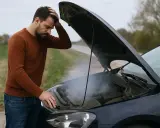 Uomo disperato con cofano aperto, fumo denso e puzza di bruciato macchina. Problema meccanico sul ciglio della strada.