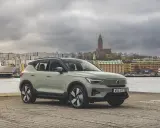 A light green Volvo XC40 parked by a waterfront with a ferry and city buildings in the background.