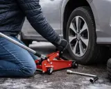 Persona che usa un cric rosso per sollevare un'auto, mostrando dove mettere il cric sotto la macchina per cambiare una gomma.