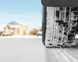 Un treno di gomme coperto di neve affronta una strada innevata, con case sfocate sullo sfondo.