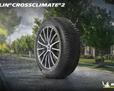 Pneumatico Michelin CrossClimate 2 in primo piano su strada bagnata, con case e alberi sullo sfondo. Nessun difetto visibile.