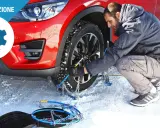 Uomo monta catene da neve su una ruota di un'auto rossa in un paesaggio innevato. Le gomme invernali sono essenziali per la sicurezza.