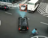 Auto rileva ciclista e attiva la forza frenante. Un'auto argentata è inquadrata, mentre un camion bianco e un'auto nera proseguono.