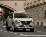 Nissan X-Trail full hybrid beige, in corsa su strada urbana con edifici storici sullo sfondo.