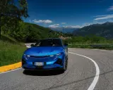 L'Alpine A290 sfreccia su una strada di montagna panoramica, con un cielo azzurro e montagne sullo sfondo.