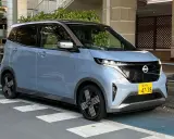 Nissan Sakura azzurro pastello, un'auto elettrica compatta con dettagli color rame sul tetto, parcheggiata su strisce pedonali.