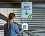 Donna interagisce con un terminale per pagare la ricarica dell'auto elettrica. Il cartello indica "Qui puoi pagare la ricarica", suggerendo il costo ricarica auto elettrica.