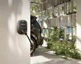 Wallbox per auto elettriche, con cavo collegato a un'auto scura. L'ambiente è moderno, con elementi verdi e pannelli solari visibili.
