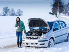 Donna al telefono accanto a un'auto d'argento con il cofano aperto, bloccata nella neve. La macchina fa fatica ad accendersi a freddo.