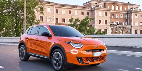Auto elettrica arancione in movimento, con 400 km di autonomia, su strada urbana con edifici storici sullo sfondo.