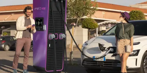 Coppia in una stazione di ricarica Enel X Way. Lei attende mentre l'auto si carica, lui controlla lo smartphone. Ottime recensioni per questa stazione di ricarica.