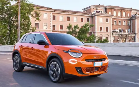 Auto elettrica arancione in movimento, con 400 km di autonomia, su strada urbana con edifici storici sullo sfondo.