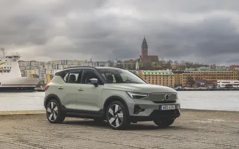 A light green Volvo XC40 parked by a waterfront with a ferry and city buildings in the background.