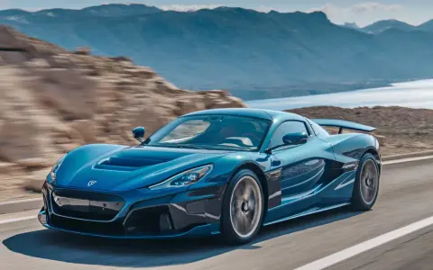 La Rimac Nevera blu sfreccia su una strada panoramica, con montagne e acqua sullo sfondo.