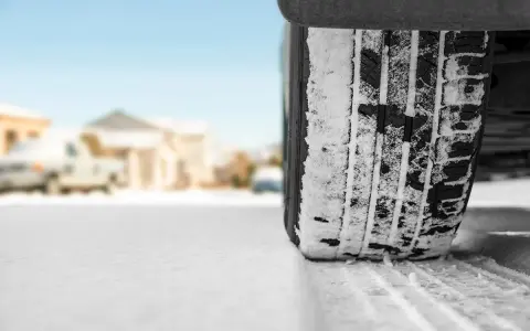 Un treno di gomme coperto di neve affronta una strada innevata, con case sfocate sullo sfondo.