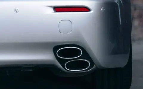 Doppio scarico cromato su un'auto sportiva argentata, parte del collettore di scarico. Sensori di parcheggio e fanale posteriore rosso visibili.