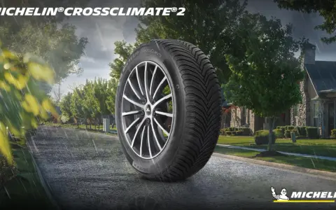 Pneumatico Michelin CrossClimate 2 in primo piano su strada bagnata, con case e alberi sullo sfondo. Nessun difetto visibile.