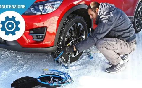 Uomo monta catene da neve su una ruota di un'auto rossa in un paesaggio innevato. Le gomme invernali sono essenziali per la sicurezza.