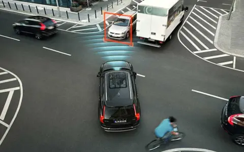 Auto rileva ciclista e attiva la forza frenante. Un'auto argentata è inquadrata, mentre un camion bianco e un'auto nera proseguono.
