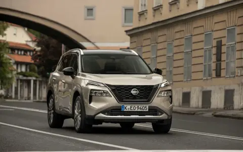 Nissan X-Trail full hybrid beige, in corsa su strada urbana con edifici storici sullo sfondo.