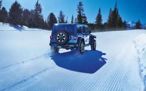 Un blu Jeep Wrangler affronta la neve con pneumatici Toyo Open Country AT3, lasciando tracce su un sentiero battuto tra alberi innevati e montagne.