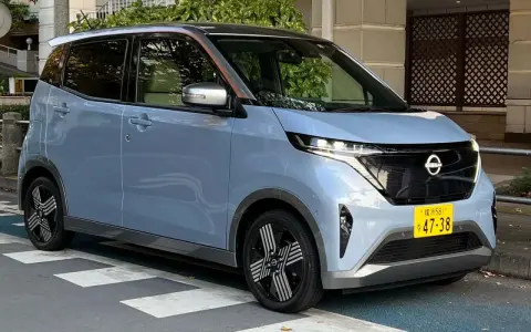 Nissan Sakura azzurro pastello, un'auto elettrica compatta con dettagli color rame sul tetto, parcheggiata su strisce pedonali.