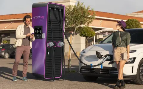 Coppia in una stazione di ricarica Enel X Way. Lei attende mentre l'auto si carica, lui controlla lo smartphone. Ottime recensioni per questa stazione di ricarica.