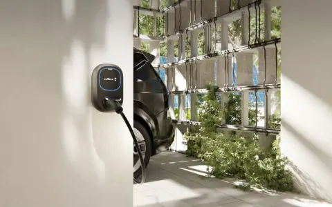 Wallbox per auto elettriche, con cavo collegato a un'auto scura. L'ambiente è moderno, con elementi verdi e pannelli solari visibili.