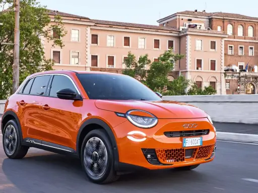 Auto elettrica arancione in movimento, con 400 km di autonomia, su strada urbana con edifici storici sullo sfondo.