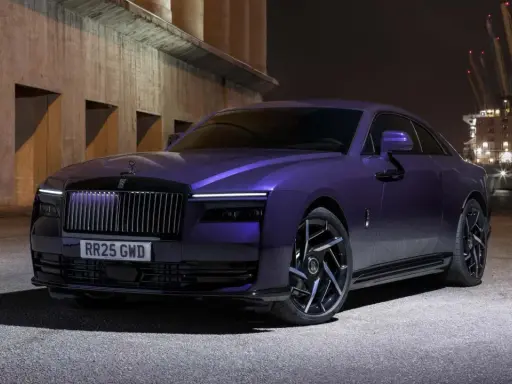 Una Rolls-Royce sportiva viola metallizzato, con un design audace e cerchi neri, parcheggiata di notte vicino a un porto industriale.