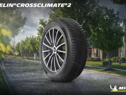 Pneumatico Michelin CrossClimate 2 in primo piano su strada bagnata, con case e alberi sullo sfondo. Nessun difetto visibile.