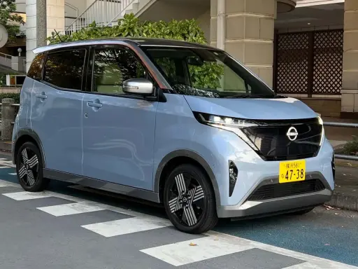 Nissan Sakura azzurro pastello, un'auto elettrica compatta con dettagli color rame sul tetto, parcheggiata su strisce pedonali.
