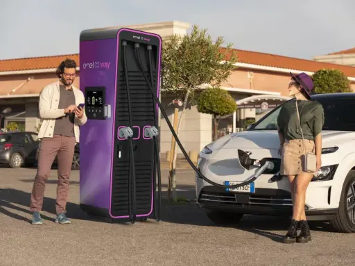 Coppia in una stazione di ricarica Enel X Way. Lei attende mentre l'auto si carica, lui controlla lo smartphone. Ottime recensioni per questa stazione di ricarica.
