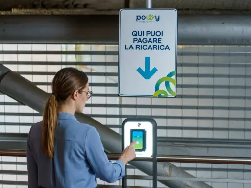 Donna interagisce con un terminale per pagare la ricarica dell'auto elettrica. Il cartello indica "Qui puoi pagare la ricarica", suggerendo il costo ricarica auto elettrica.