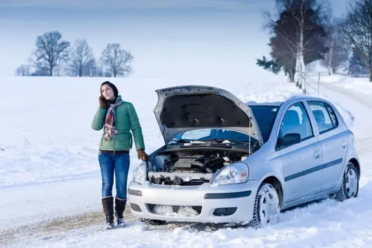 Donna al telefono accanto a un'auto d'argento con il cofano aperto, bloccata nella neve. La macchina fa fatica ad accendersi a freddo.