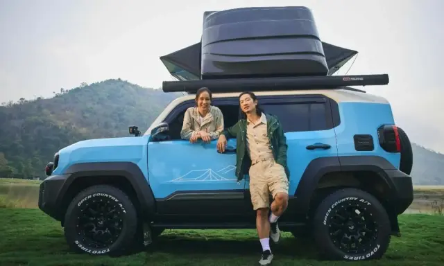 Coppia sorridente accanto a un toyota land cruiser mini blu, pronto per l'avventura con tettoia e box sul tetto.