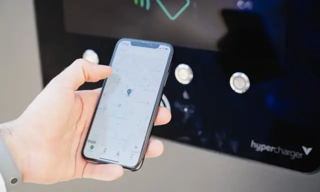 Mano che usa smartphone con mappa per trovare stazione di ricarica. L'app mostra la posizione e le opzioni per monitorare la carica.