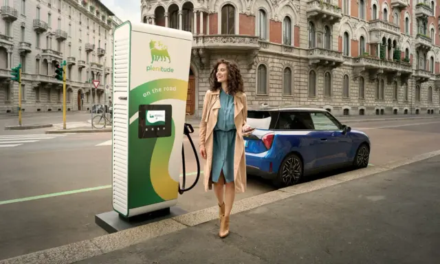 Donna sorridente vicino a colonnina di ricarica EVS ENI e auto elettrica blu, su strada cittadina con palazzi storici.