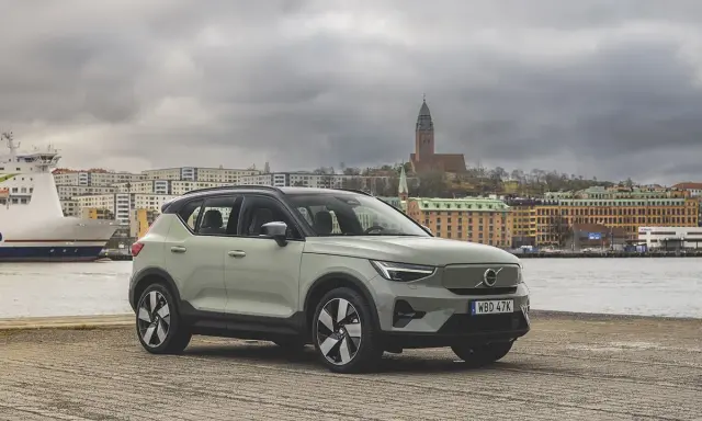 A light green Volvo XC40 parked by a waterfront with a ferry and city buildings in the background.