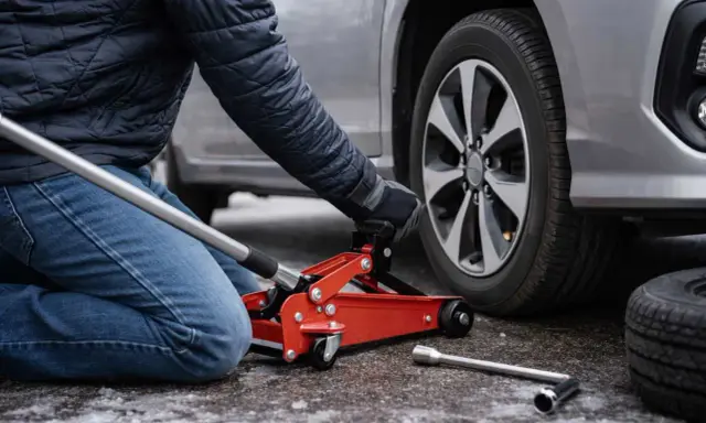 Persona che usa un cric rosso per sollevare un'auto, mostrando dove mettere il cric sotto la macchina per cambiare una gomma.